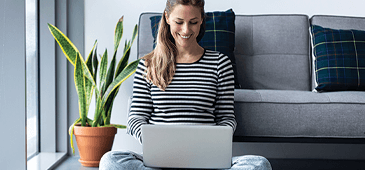 smiling person using a laptop sitting against a sofa