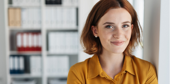 Person in a mustard-yellow shirt standing indoors in front of shelves with binders, facing the camera in a bright office setting.